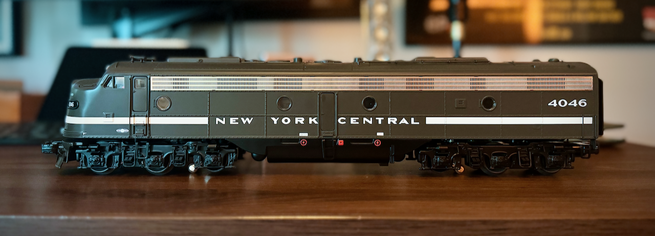 A side view of a model EMD E8 diesel locomotive in New York Central livery, sitting on a wooden surface. The streamlined body is painted very dark grey, almost black, with thin white pinstriping along the lower edge; 'NEW YORK CENTRAL' is lettered in white across the side and the cab number '4046' appears beneath the engineer's window. A row of small porthole-style windows runs along the carbody. The background is softly out of focus, keeping attention on the model.