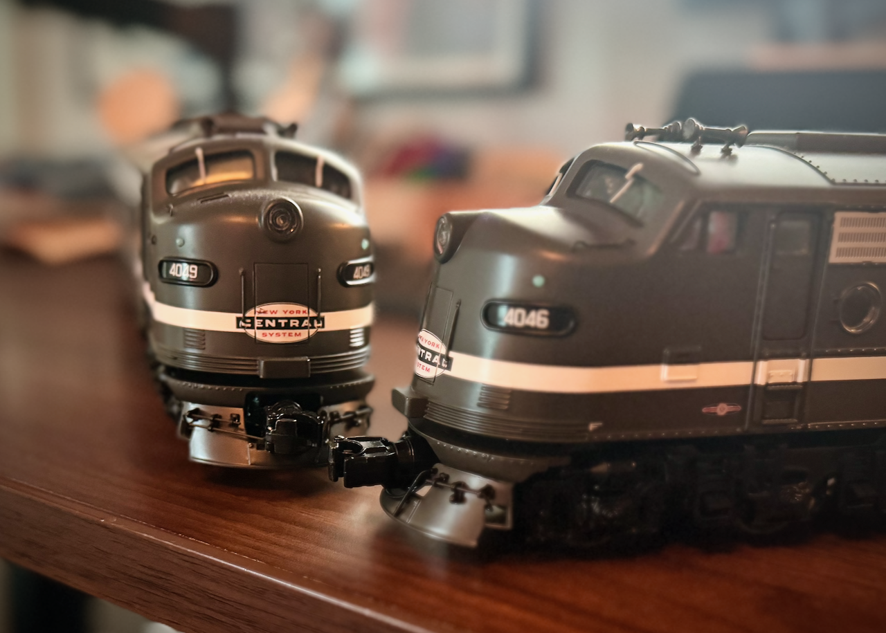 The image shows two model train locomotives placed on a wooden surface. They are painted in a dark green color with white stripes and have the 'New York Central System' logo prominently displayed on their fronts. The locomotive on the left has the number '4049' and the one on the right has the number '4046.' Both locomotives have a streamlined design typical of mid-20th century diesel engines. The background is slightly blurred, keeping the focus on the two model trains.