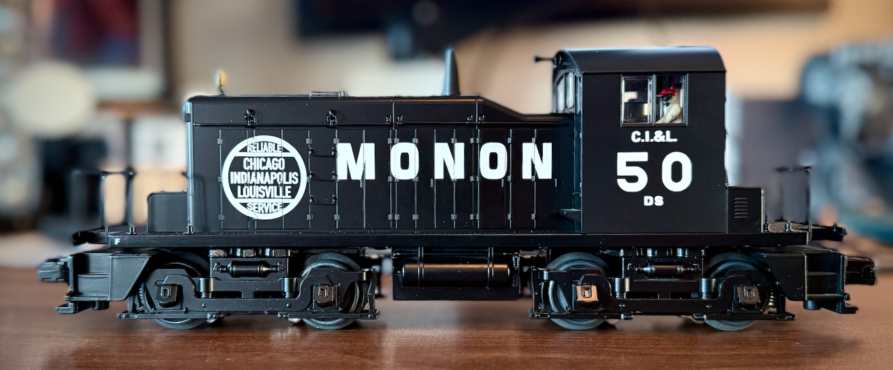 This image shows a detailed model of the Monon DS-50 locomotive. The black engine features bold white lettering reading 'MONON' on the side, along with a circular emblem that says, 'Reliable Chicago Indianapolis Louisville Service.' The number '50' and is visible on the cab. The model is set on a table, with a softly blurred background, highlighting the craftsmanship of this vintage railroad replica.