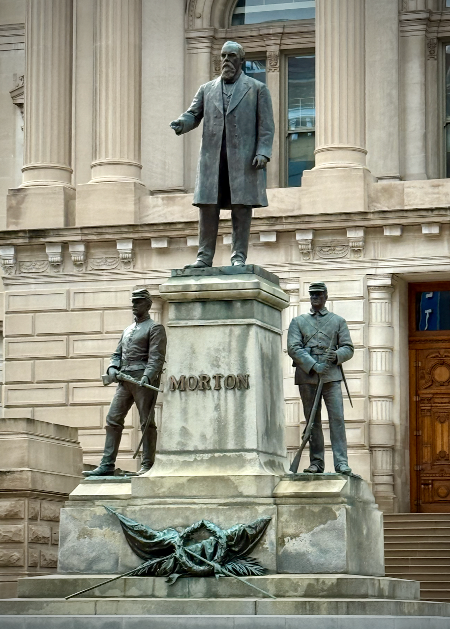 The image shows a statue of Oliver P. Morton, positioned in front of the Indiana Statehouse. Morton, a prominent figure in Indiana's Civil War history, stands at the top of the monument, with two soldiers depicted on either side at the base. The soldiers, dressed in Civil War uniforms, representing the Union, reflecting Morton's role during the war. The pedestal bears Morton's name, with a decorative laurel wreath and flags at the bottom.
